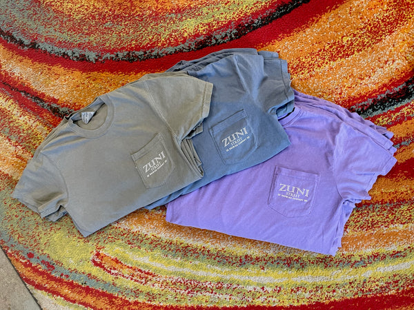 Three folded shirts in gray, blue, and purple on a colorful woven surface with 'ZUNI' branding.
